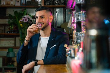 Handsome Caucasian man sitting at bar counter holding whiskey glass with ice cube and drinking alcoholic drink from barman in nightclub. Nightlife holiday party celebration and small business concept