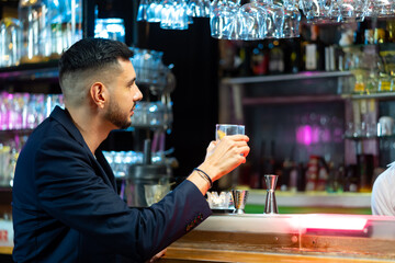 Handsome Caucasian man sitting at bar counter holding whiskey glass with ice cube and drinking alcoholic drink from barman in nightclub. Nightlife holiday party celebration and small business concept