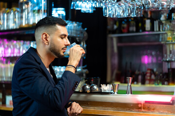 Handsome Caucasian man sitting at bar counter holding whiskey glass with ice cube and drinking alcoholic drink from barman in nightclub. Nightlife holiday party celebration and small business concept