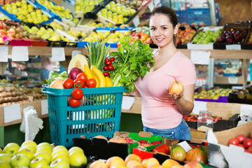 Young satisfied woman with basket filled with fresh fruits and vegetables in the store