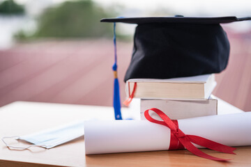 Closed-up photo of a college graduation cap mortarboard with degree diploma certificate and face mask on the table. Graduation in the era of COVID-19