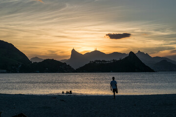 Surfista observa mar calmo e crianças brincando na agua ao pôr-do-sol na praia 