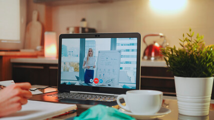 Online video conference on laptop with female speaker in home office. In the foreground: writing hands, medical mask and a cup.