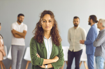 Portrait of a young serious business woman standing in an office hall against the backdrop of employees. Beautiful curly woman stands with folded arms and looks at the camera. Confidence concept.