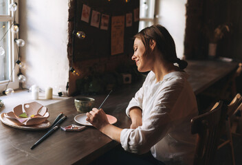 Young attractive woman in white shirt ceramic artist decorating clay plate with tool at table in pottery workshop. Handmade work student, freelance small business