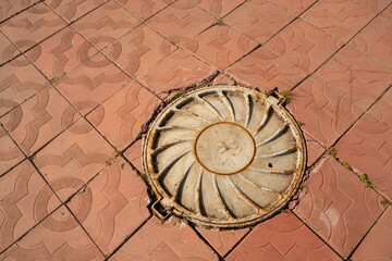 Embossed cast-iron cover of the manhole - part of the manhole cover is located on the pavement made of concrete slabs.
