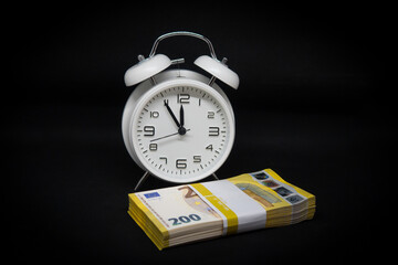 bundle of 200-euro banknotes is lying in front of a white alarm clock with its hands set to 5 minutes to 12