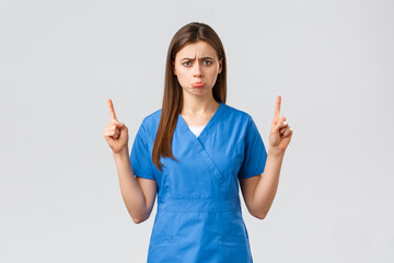 Healthcare workers, prevent virus, insurance and medicine concept. Upset sobbing cute nurse or doctor in blue scrubs, pouting, feel uneasy pointing fingers up at unpleasant bad news, show banner