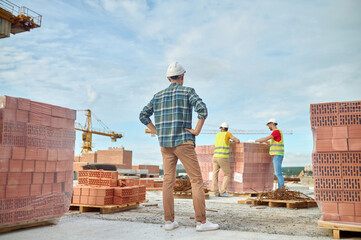 Foreman watching workers leaning on the stack of red bricks