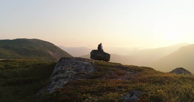Hiker sitting alone on mountain rock enjoying Lofoten sunset - aerial orbit
