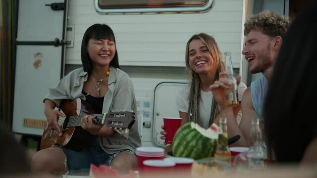 Smiling friends are enjoying guitar songs while having a picnic sitting near the caravan trailer outside