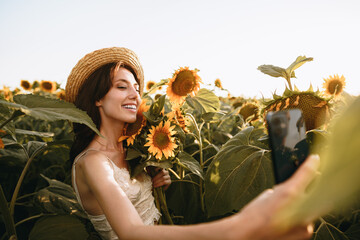 Young smiling woman in hat taking selfie photo in sunflower field