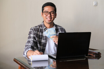 Adult Asian man smiling while holding paper money in front of laptop