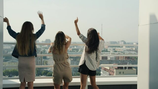 Happy young women friends are dancing having fun watching cityscape from balcony of scyscraper on summer day. Open air party and friendship concept.