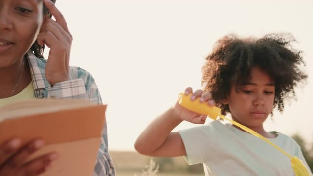 A beautiful afro-american mother is reading a book while her children blow bubbles during picnic time outside