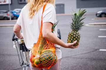 Woman with mesh bag holding pineapple in a supermarket parking lot. Female person with shopping cart after groceries purchased. Zero waste and sustainable lifestyle concept - Powered by Adobe