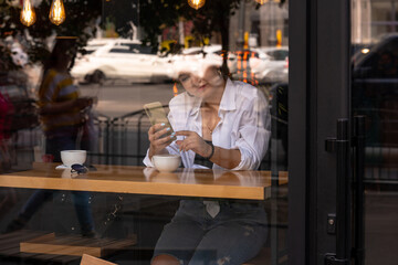 Woman at trendy cafe drinking coffee or tea and use smartphone. Young beautiful model in white shirt and red lipstick. Reflections from windows. Shot through the window of fashion girl.