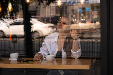 Woman at trendy cafe drinking coffee or tea. Young beautiful Caucasian model in white shirt and red lipstick. Reflections from windows. Image cross processed. Shot through the window of fashion girl.