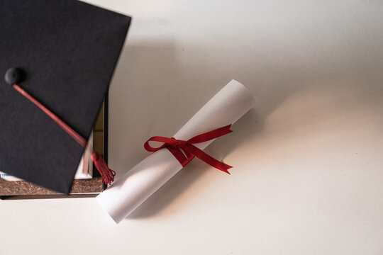 A graduation hat or mortarboard and diploma certificate paper tied with red ribbon on a stack of  books with empty space slightly undersaturated with vignette for vintage effect