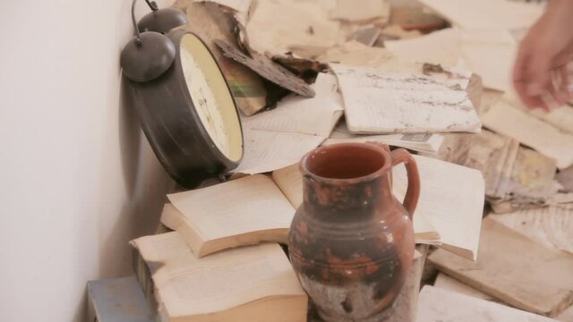 Piles of old books and a broken alarm clock inside a room, into an abandoned house. The hand of a man browsing the pages.
