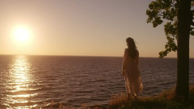Romantic young woman standing on river bank alone, looking at beautiful sunset