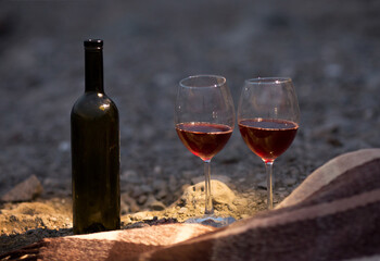 two glasses of red wine and a bottle are standing on the beach next to a blanket