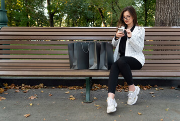 Young woman with phone and shopping bags in park. Online shopping. Discounts, promotions, black friday