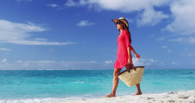 Woman tourist walking on tropical summer vacation wearing sun hat, red dress and beach bag relaxing on travel holidays. Young lady in luxury fashion beachwear. SLOW MOTION VIDEO