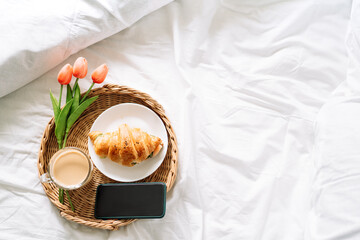 Croissant and coffee on white bed on wicker tray with flowers