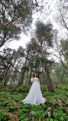 A bride in a white dress stands at the trees in the mountains