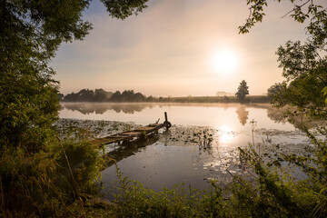 Naklejka na ściany i meble Fishing bridge for a boat and a calm morning landscape surrounded by greenery at sunrise with fog over the river.