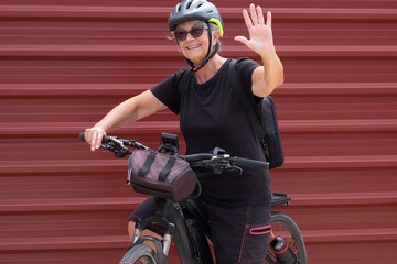 Fototapeta premium Happy senior woman cyclist in outdoor excursion close to a red metal panel. Elderly people ejoying healthy lifestyle