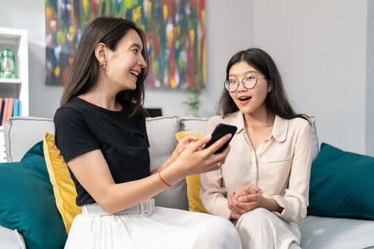 Beautiful Asian woman sits on the couch with a surprised friend, shows her messages sent on social media, photos on the phone screen, the girl makes a shocked face, opens her mouth