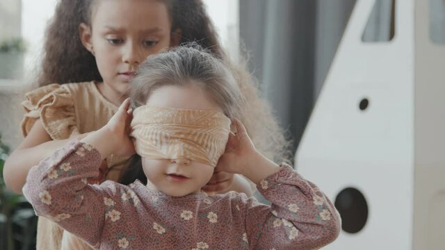 Slowmo medium shot of 8-year-old girl with curly hair putting blindfold on cute little girl while playing tag at home