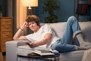 Obraz premium A young boy with curly hair lies on the couch at home watching TV with wireless headphones. Teenager with a smile listens to music,podcast,news while relaxing in the evening
