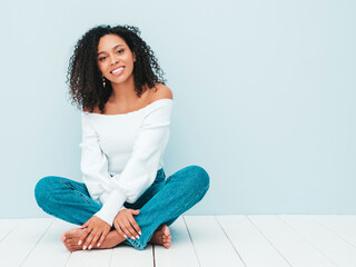 Beautiful black woman with afro curls hairstyle.Smiling model in sweater and trendy jeans clothes. Sexy carefree female sitting near light blue wall in studio. Tanned and cheerful