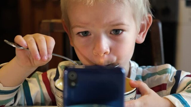 Little Boy using Phone while eating Soup during Lunch or Dinner at the Table. Child eating Food and watch Cartoon on Smartphone because Nomophobia or Attention Deficit Hyperactivity Disorder ADHD .