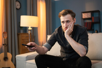 Bored guy sits on the couch in the evening in living room, holding the TV remote in his hand,...