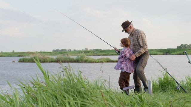 Fishing for pike, perch, carp. Skilled senior Fisherman and child boy with rod, spinning reel on river bank. Man and child catching fish, pulling rod while fishing on lake, happy friendly family