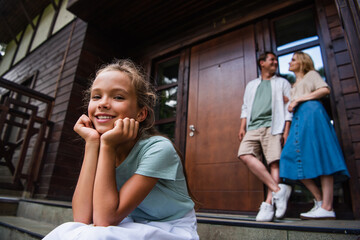 Girl looking at camera on stairs near blurred parents and vacation house