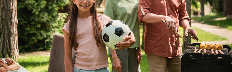 Cropped view of kid holding football ball near parents cooking on grill, banner