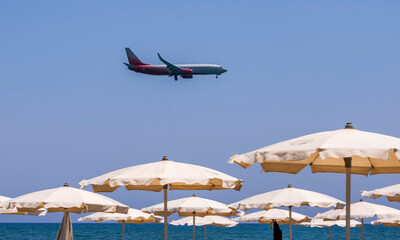 Fototapeta na wymiar passenger plane flying low above beach with blue sea with resting and swiming tourists and parasol umbrellas on the foreground