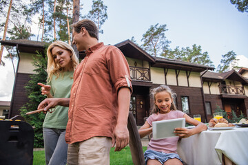 Low angle view of smiling kid using digital tablet near parents cooking on grill