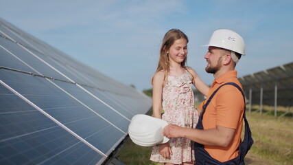A father, a solar power engineer, and his daughter are standing near solar panels. The father explains to the child the principle of solar electricity and puts a protective helmet on the girl's head.