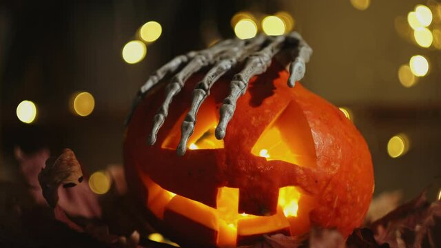 Skeleton hand holds Halloween pumpkin head illuminated from inside. Scary carved pumpkin Jack o lantern. Halloween home decor.