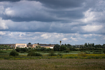 athmospheric moody picture of cloudy sky with dark rainy or stormy clouds, green meadow or pasture with some bush and old factory buildings in distance. Latvian common flat landscape