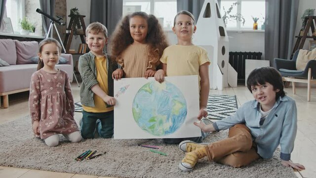Portrait of group of kids holding drawing of planet Earth and rocket ship and posing for camera in living room