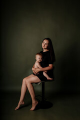 Fototapeta na wymiar a brunette mother in a black dress is sitting on a chair with her baby daughter on a green isolated background with a place for text