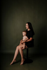 beautiful mother in a black dress is sitting on a chair and playing with her baby daughter on a green isolated background with a place for text