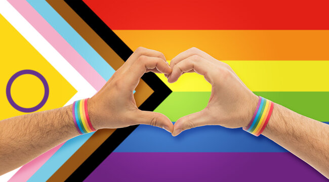 lgbtq, trans and intersex rights concept - close up of male couple hands with rainbow wristbands showing heart gesture over progress pride flag on background